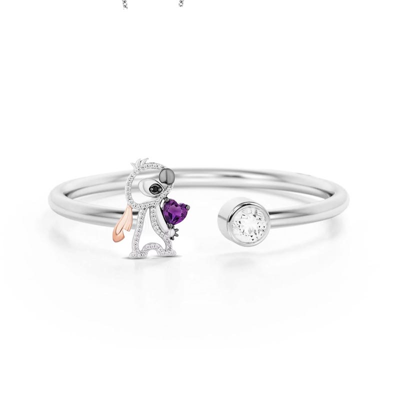May include: A silver-toned open cuff bracelet featuring a stylized character holding a purple heart-shaped gem. The character has a silver body, black eyes, and a rose gold ear. A clear, round gem is set on the opposite end.