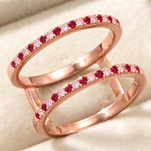 Puede incluir: Un anillo de doble banda en oro rosa con dos filas paralelas de piedras preciosas redondas que alternan entre rojo y rosa claro. El metal tiene un acabado pulido y las piedras están engastadas a lo largo de la mitad superior de cada banda.