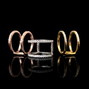 Puede incluir: Tres anillos se muestran sobre un fondo negro. Un anillo es de tono plateado con dos bandas horizontales conectadas por barras verticales, todas incrustadas con pequeñas piedras claras. Otros dos anillos son bandas de estilo abierto con piedras claras.