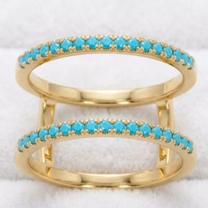 Puede incluir: Un anillo de doble banda hecho de metal dorado, con dos bandas paralelas conectadas por barras verticales. Cada banda está incrustada con una fila continua de pequeñas piedras redondas de color turquesa engastadas en estilo pavé.