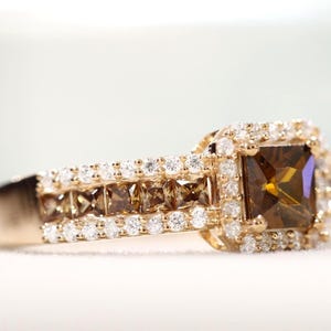 Può includere: Un anello d'oro con una grande pietra marrone a taglio quadrato al centro, circondata da pietre bianche e marroni più piccole. La fascia è inoltre decorata con molte piccole pietre preziose bianche rotonde.