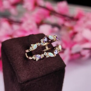 Op de afbeelding: Een goudkleurige ring met twee banden, versierd met witte en paarse edelstenen. De edelstenen zijn gerangschikt in een bladvormig patroon, met kleine heldere stenen ertussen. De ring staat tegen een donkerbruine achtergrond met een vage roze bloemenachtergrond.