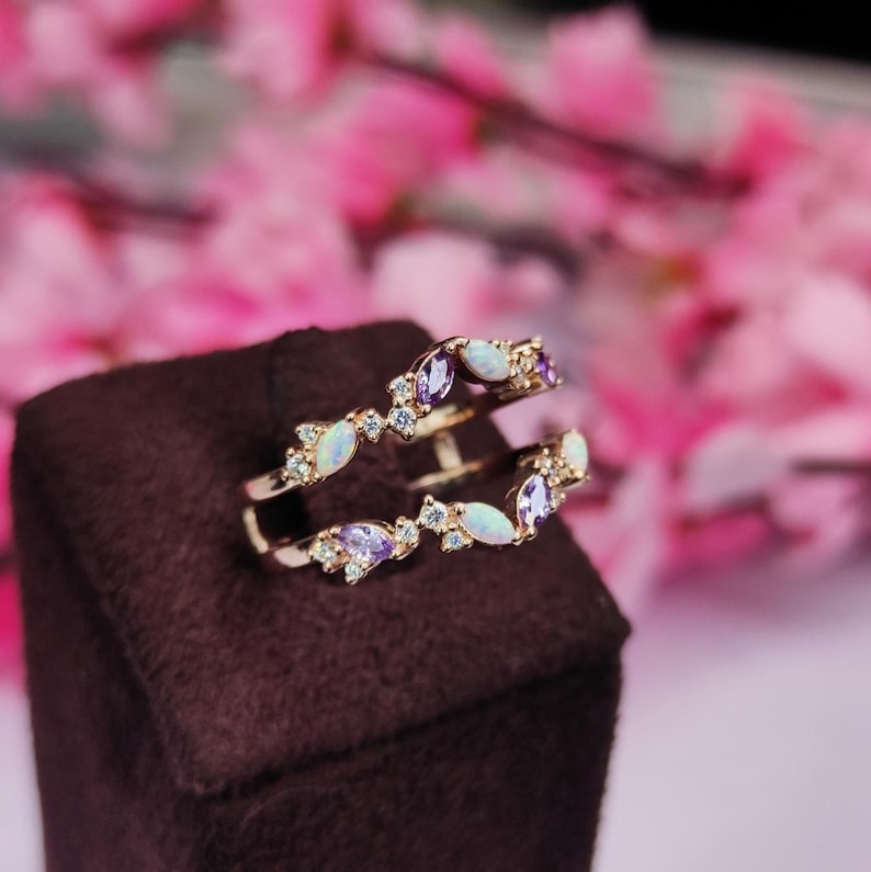 Op de afbeelding: Een goudkleurige ring met twee banden, versierd met witte en paarse edelstenen. De edelstenen zijn gerangschikt in een bladvormig patroon, met kleine heldere stenen ertussen. De ring staat tegen een donkerbruine achtergrond met een vage roze bloemenachtergrond.