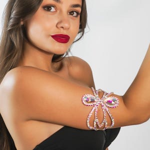 May include: A silver-toned arm cuff with pink and clear crystals. The cuff features a bow-like design with dangling crystal accents. The jewelry is worn on the upper arm. The background is plain white.