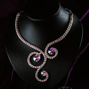 Puede incluir: Un collar de cristal blanco con un dise&ntilde;o de espiral y piedras iridiscentes. El collar se muestra en un soporte de terciopelo negro.