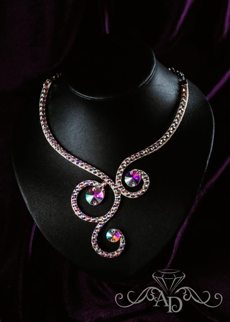 Puede incluir: Un collar de cristal blanco con un dise&ntilde;o de espiral y piedras iridiscentes. El collar se muestra en un soporte de terciopelo negro.