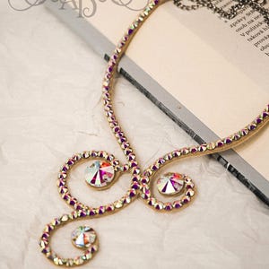 Puede incluir: Un collar de oro con un dise&ntilde;o delicado que presenta m&uacute;ltiples espirales y brillantes diamantes de imitaci&oacute;n iridiscentes. El collar est&aacute; sobre una superficie blanca.