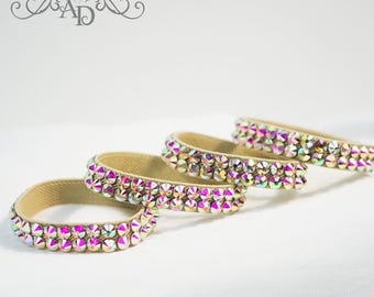 Puede incluir: Cuatro pulseras de pedrería iridiscentes. Cada pulsera tiene una banda de tela beige con dos filas de pedrería colorida. Las pulseras están dispuestas en diagonal sobre una superficie blanca, con un logotipo de diamante en la esquina superior izquierda.