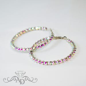 Può includere: Un paio di orecchini a cerchio con cristalli iridescenti multicolori. I cristalli sono disposti attorno a un cerchio di metallo argentato. Gli orecchini sono esposti su sfondo bianco. Gli orecchini hanno una chiusura.