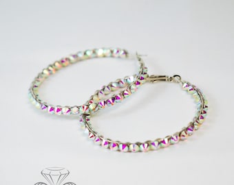 May include: A pair of hoop earrings with iridescent, multi-colored crystals. The crystals are arranged around a silver-toned metal hoop. The earrings are displayed on a white background. The earrings have a clasp closure.