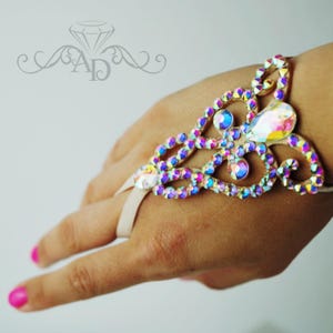 Puede incluir: Un brazalete de mano adornado con cristales iridiscentes en tonos morados, azules y amarillos. El diseño incluye un gran cristal central y piedras más pequeñas, conectadas por una delicada banda de color claro que rodea el dedo.