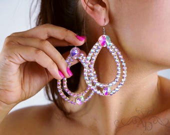Puede incluir: Grandes pendientes iridiscentes en forma de lágrima con múltiples capas de cristales brillantes. Los pendientes se sostienen cerca de la oreja, mostrando su tamaño y la forma en que capturan la luz. Los cristales reflejan un arcoíris de colores.