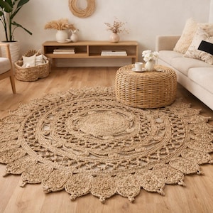 May include: A round, natural jute rug with an intricate, floral-inspired design. The rug is beige and tan, with a detailed pattern in the center and a scalloped edge. It is placed on a light wood floor, with a sofa and chair in the background.