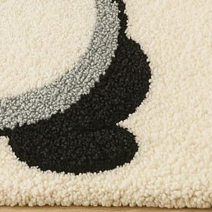May include: Close-up of a cream-colored, textured rug with a large, abstract design. The design features a curved shape in black and gray, creating a bold contrast against the neutral background. The rug's texture suggests a soft, plush feel.
