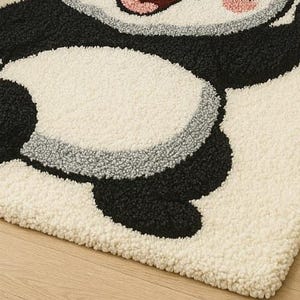 May include: A cream-colored, plush rug featuring a playful panda design. The panda has black and white fur with pink accents on its paws and face. The rug's texture is soft and inviting, perfect for a child's room or a whimsical space.