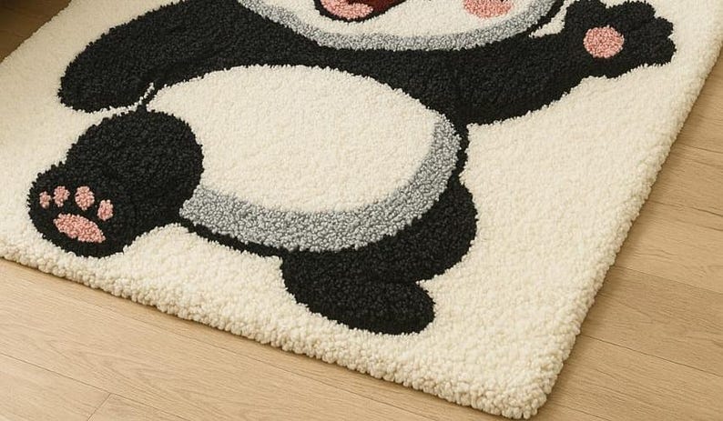 May include: A cream-colored, plush rug featuring a playful panda design. The panda has black and white fur with pink accents on its paws and face. The rug's texture is soft and inviting, perfect for a child's room or a whimsical space.