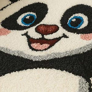 May include: A close-up of a panda rug with a smiling face. The rug features a black and white design with blue eyes, a red nose, and pink accents. The rug is made of a soft, textured material.