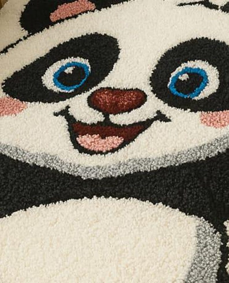 May include: A close-up of a panda rug with a smiling face. The rug features a black and white design with blue eyes, a red nose, and pink accents. The rug is made of a soft, textured material.
