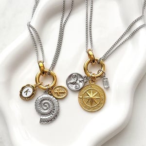 Puede incluir: Dos collares de cadena plateada con anillos circulares dorados como colgantes. El primer collar incluye un dije ovalado blanco con la letra K, un dije de fósil de amonita plateado y una pequeña cruz dorada. El segundo collar presenta un dije del zodiaco plateado, un dije de brújula dorada y un pequeño dije de cristal rectangular.
