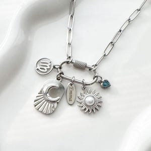 Op de afbeelding: Een zilverkleurige ketting met een karabijnsluiting versierd met meerdere bedels: een Maagd sterrenbeeld symbool, een rechthoekig plaatje met een halve maan en zon, een ovaal plaatje met de inscriptie Emma, een zon met een parel en een klein blauw hartje.