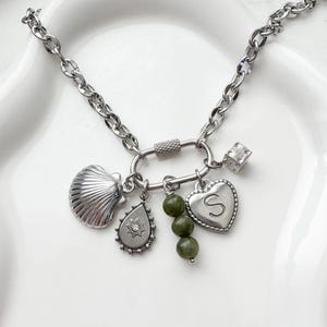 May include: A silver necklace featuring multiple charms. The charms include a seashell, a teardrop with a starburst design, a heart with the letter "S", a dice, and green beads. The necklace has a chain link design and a clasp.