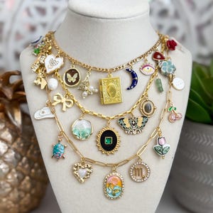 Puede incluir: Un collar de cadena dorada con múltiples dijes, incluyendo una mariposa, una luna creciente, un corazón, una tortuga, una flor, un libro, una gema, una cereza, un tulipán y un signo del zodíaco. Los dijes están todos esmaltados en varios colores, incluyendo verde, azul, rojo, rosa y blanco.