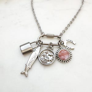May include: A silver-toned chain necklace featuring a carabiner clasp as a pendant holder. Attached to the carabiner are four charms: a rectangular padlock, a fish, a circular medallion with a scorpion and star, and a sunburst charm with a pink stone.