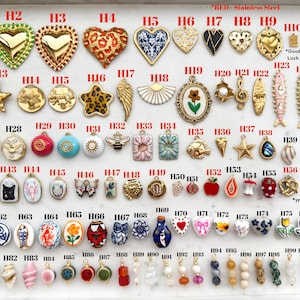 May include: A collection of assorted metal, enamel, and ceramic charms arranged in rows on a white tray. The charms feature various shapes including hearts, stars, animals, flowers, and geometric patterns, each labeled with an alphanumeric code.