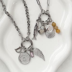 May include: Two silver-toned chain necklaces featuring circular clasps with multiple charms. One necklace includes a letter D, a circular sun medallion, a pink rectangular crystal, and a wing. The other features a fish, heart, moon, and yellow beads.
