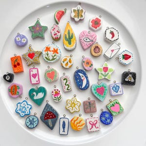 May include: A collection of colorful, hand-painted charms in various shapes, including stars, hearts, and fruits. Each charm features unique designs, such as flowers, cacti, and abstract patterns. The charms are arranged on a white surface.