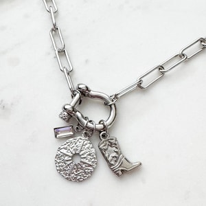 May include: A silver-toned chain necklace featuring a horseshoe-shaped clasp. Attached to the clasp are three charms: a rectangular iridescent crystal, a textured circular disc with a central hole, and a small western-style cowboy boot.