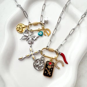 May include: Two necklaces with silver-tone chains and various charms. Charms include a butterfly, cross, dice, horseshoe, and a red chilli pepper. The necklaces are displayed on a white surface.
