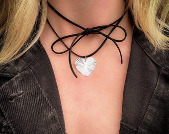 May include: A silver heart pendant necklace with a black cord choker. The heart pendant has a textured, radial design. The cord is tied in a bow at the back of the neck.