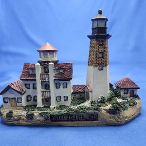 May include: A miniature lighthouse figurine of Point Judith, RI, featuring a tall tower with a brick-patterned top and white base, attached to a multi-story white building with red shingled roofs, all set upon a textured rocky base.