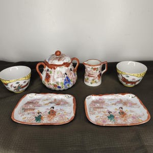 May include: A vintage Japanese porcelain tea set featuring a sugar bowl with lid, creamer, two teacups, and two small rectangular plates. The set is decorated with colorful scenes of figures and landscapes, with red and gold trim.