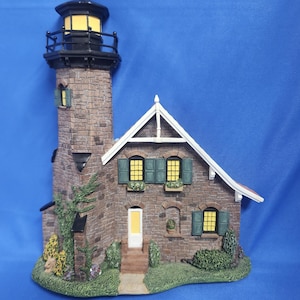 May include: A detailed miniature lighthouse with a brick tower and attached house. The lighthouse features a black lantern room and a yellow light. The house has green shutters, a white door, and a small garden. The base is surrounded by green grass and shrubbery.