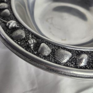 May include: A silver serving bowl with a raised rim featuring a decorative design of seashells. The bowl is made of metal and has a smooth, polished finish.