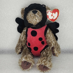 May include: A plush teddy bear wearing a black hood, black wings, and a red ladybug-themed outfit with black spots. The bear has a Ty heart-shaped tag attached.