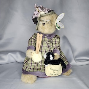 May include: A plush brown bear wearing a purple and green plaid dress, a purple hat with purple flowers, and glasses. The bear is holding knitting needles and yarn. A black and white cat is sitting in a beige bag that says "Knitting is my bag".