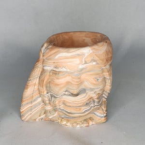 May include: A decorative ceramic planter with a unique marbled design. The planter features a blend of tan, cream, and gray tones, creating a swirling pattern. The open top is ideal for small plants or succulents. The overall shape is organic and textured.