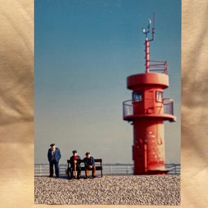 May include: Three miniature figures wearing blue jackets and hats sit on a bench in front of a red lighthouse with a white railing. The lighthouse is on a rocky shore.