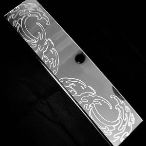 May include: A rectangular, reflective mirror surface featuring a white, etched graphic design of stylised, swirling ocean waves. The design is positioned at the top left and bottom right corners of the elongated, smooth, and metallic-looking object.