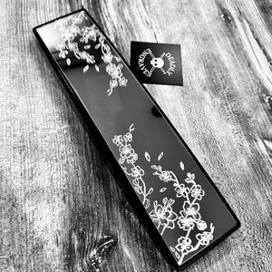 May include: Black rectangular mirror with a white floral design. The mirror is on a wooden surface.