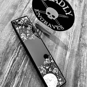 May include: A black rectangular mirror with a white design of moons, stars, and hearts. The mirror is sitting on a wooden surface. The text "DEADLY ENGRAVES" is visible on a black circle behind the mirror.