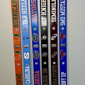 May include: A collection of six wooden decorative items, each painted in a different color and featuring college team logos and text. The teams include Miami Hurricanes, North Carolina Tar Heels, Florida Gators, UCF Knights, Florida State Seminoles, and SMU Mustangs.