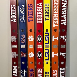 May include: A collection of seven wooden decorative items, each painted in a different color and featuring college team names and logos. The items are long and narrow, with a hole near the top and team names like "Texas A&M" and "Alabama" in white.