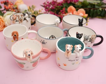 May include: A collection of ceramic mugs with 3D dog and cat figurines. The mugs are white, pink, and teal, with paw prints and strawberry designs. Some mugs have black rims and handles. The mugs are on a pink surface.