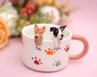 May include: A white ceramic mug with a pink handle, featuring two dog figurines inside. The mug is decorated with paw prints in red, orange, and gray. The names "Toby" and "Kajtek" are written on the mug.