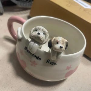 May include: White ceramic mug with a pink handle and two dog-shaped spoon rests. The spoon rests are white with brown accents and have the names "Risa" and "Bernardo" written on the mug.