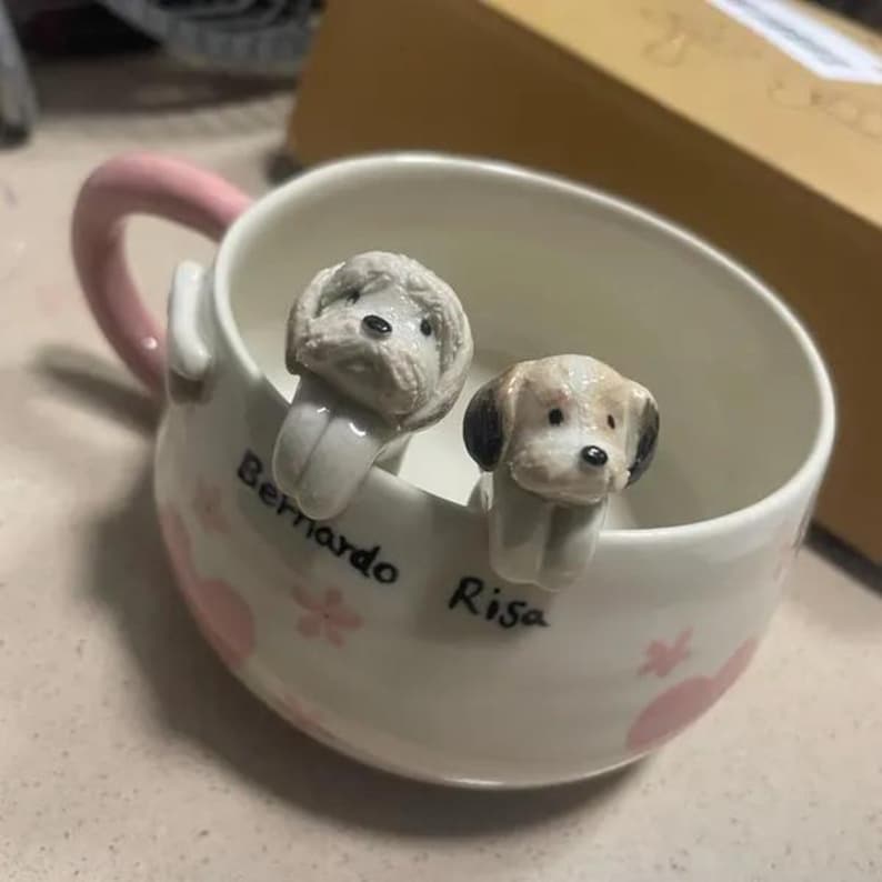 May include: White ceramic mug with a pink handle and two dog-shaped spoon rests. The spoon rests are white with brown accents and have the names "Risa" and "Bernardo" written on the mug.
